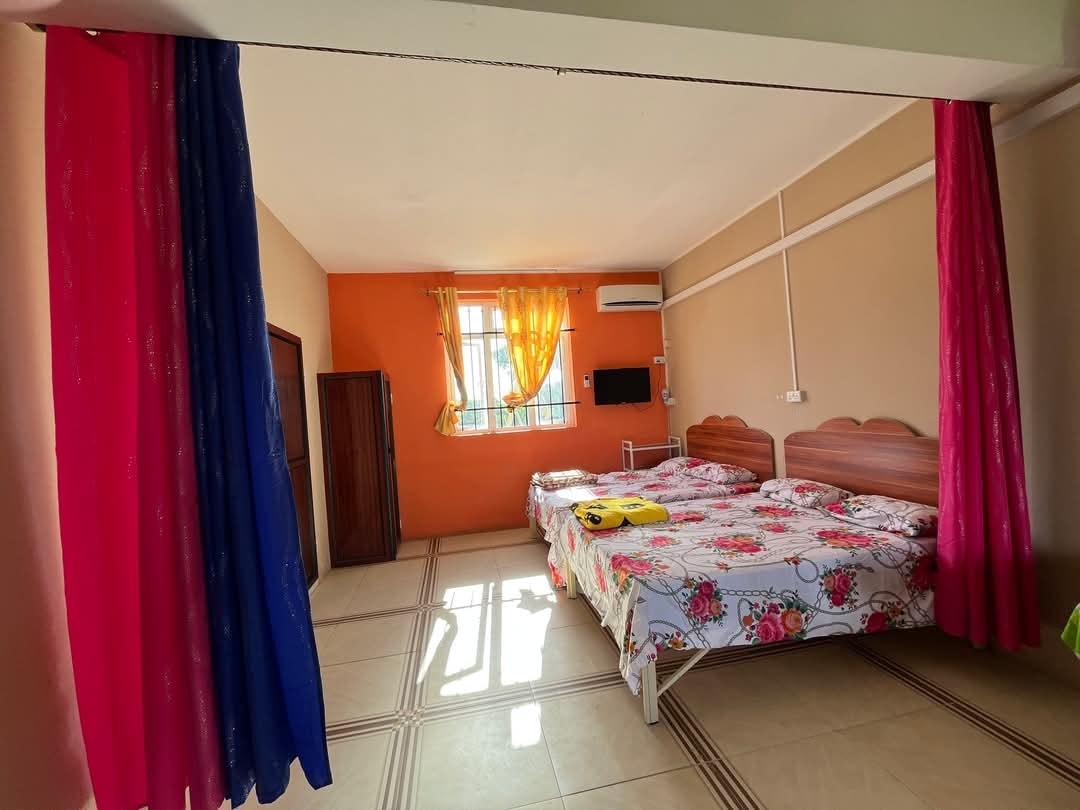 A brightly colored room features two beds with floral bedding. Sheer curtains in pink and blue frame the windows, allowing natural light to fill the space. An air conditioning unit and a wall-mounted TV are visible, with a wardrobe providing additional storage.