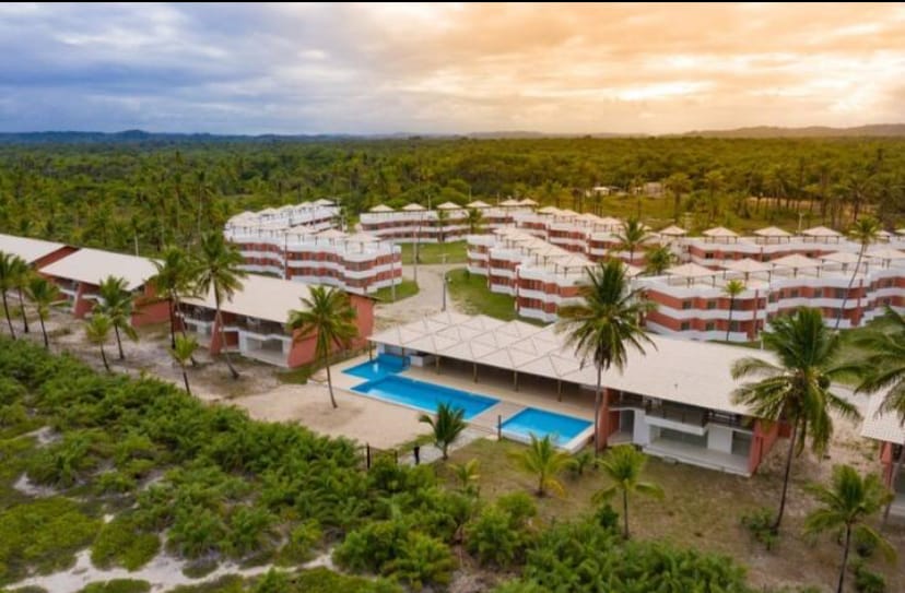 Aerial view of a serene beachfront complex featuring multiple low-rise buildings surrounded by lush greenery and palm trees. A central area includes a swimming pool, with comfortable lounge spaces under a shaded canopy. The sky reflects warm hues of orange and yellow during sunset.