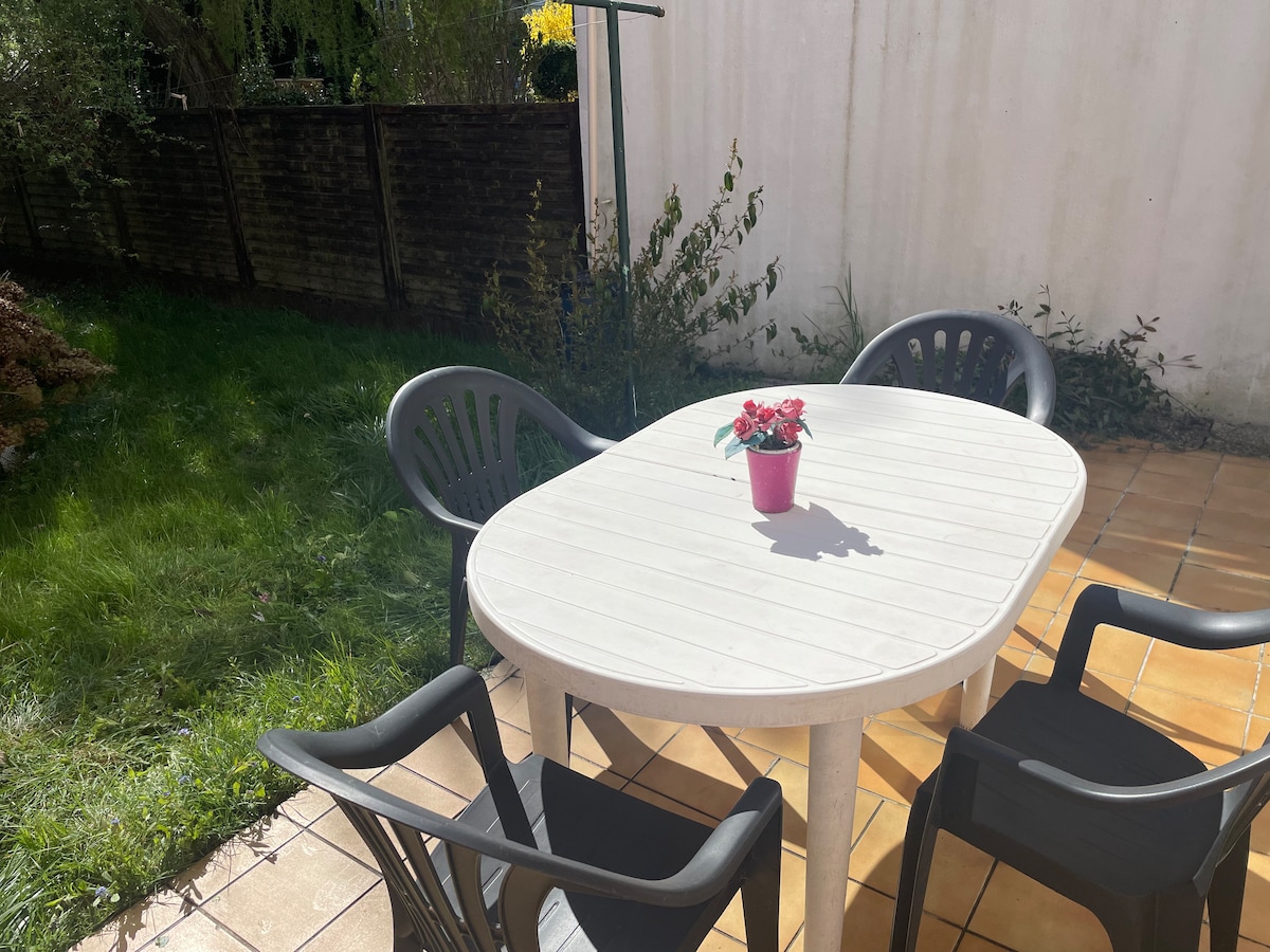 An outdoor dining area is depicted with a round table surrounded by four black chairs. A small potted plant with vibrant flowers sits at the center of the table. Soft grass and a stone-paved surface complete the inviting space, framed by a garden backdrop.