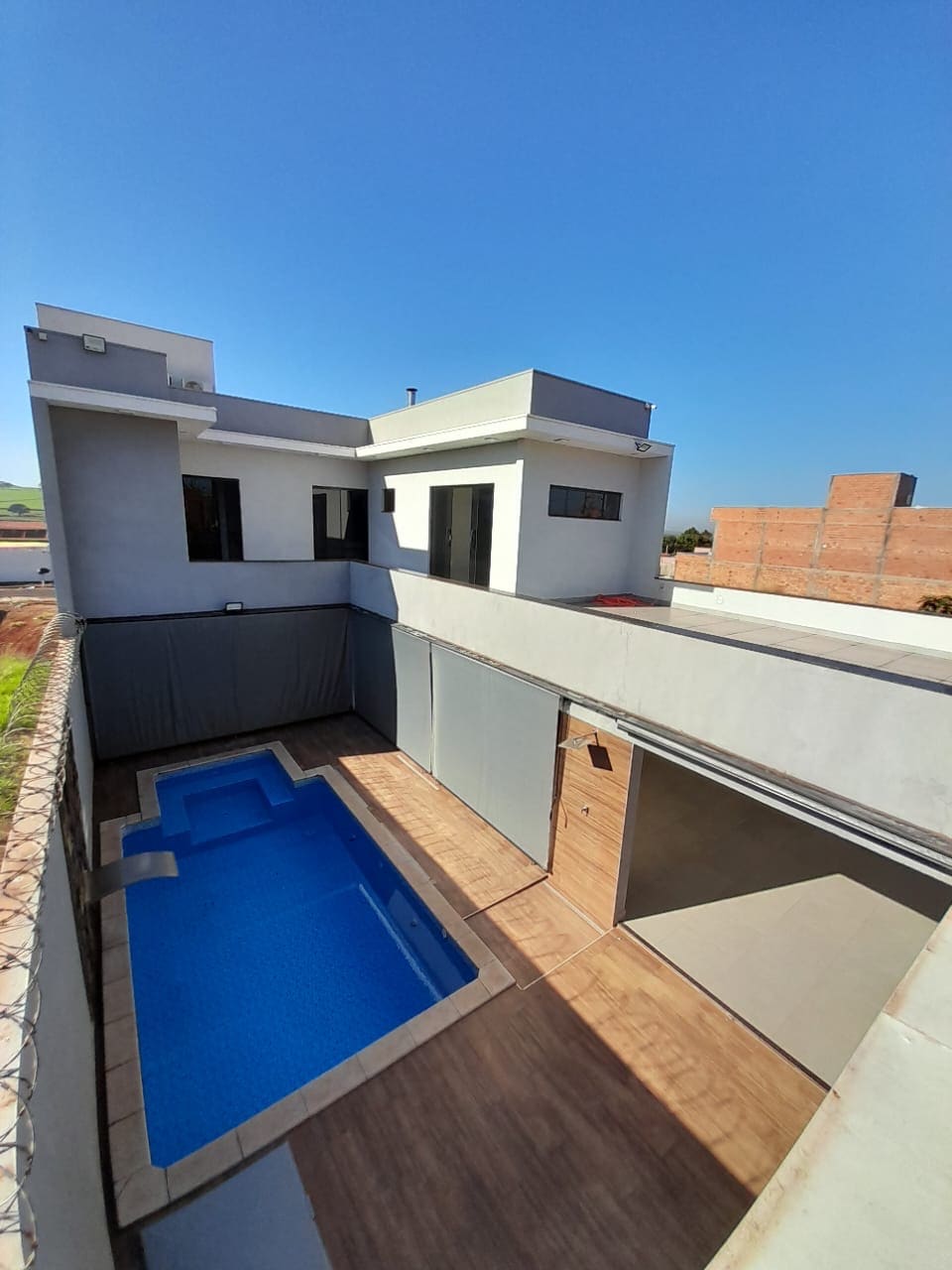An aerial view of a modern house showcases a private swimming pool bordered by a patio area. The design includes multiple levels with large windows allowing natural light. The surrounding landscape features open space and nearby structures.