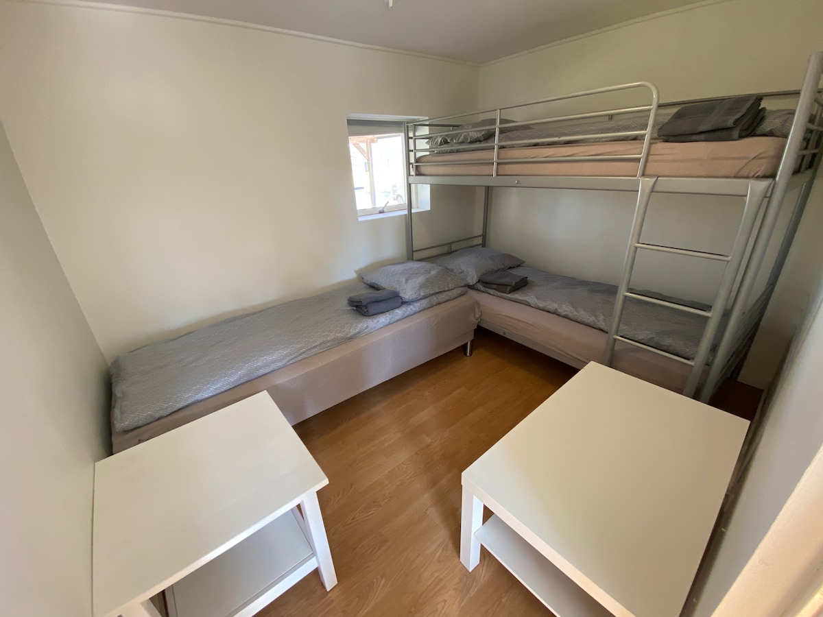A simple bedroom is presented with a bunk bed and a separate single bed. Two small pillows and a few gray blankets are neatly arranged on the beds. A white table is positioned in the center of the room, enhancing the functional layout.