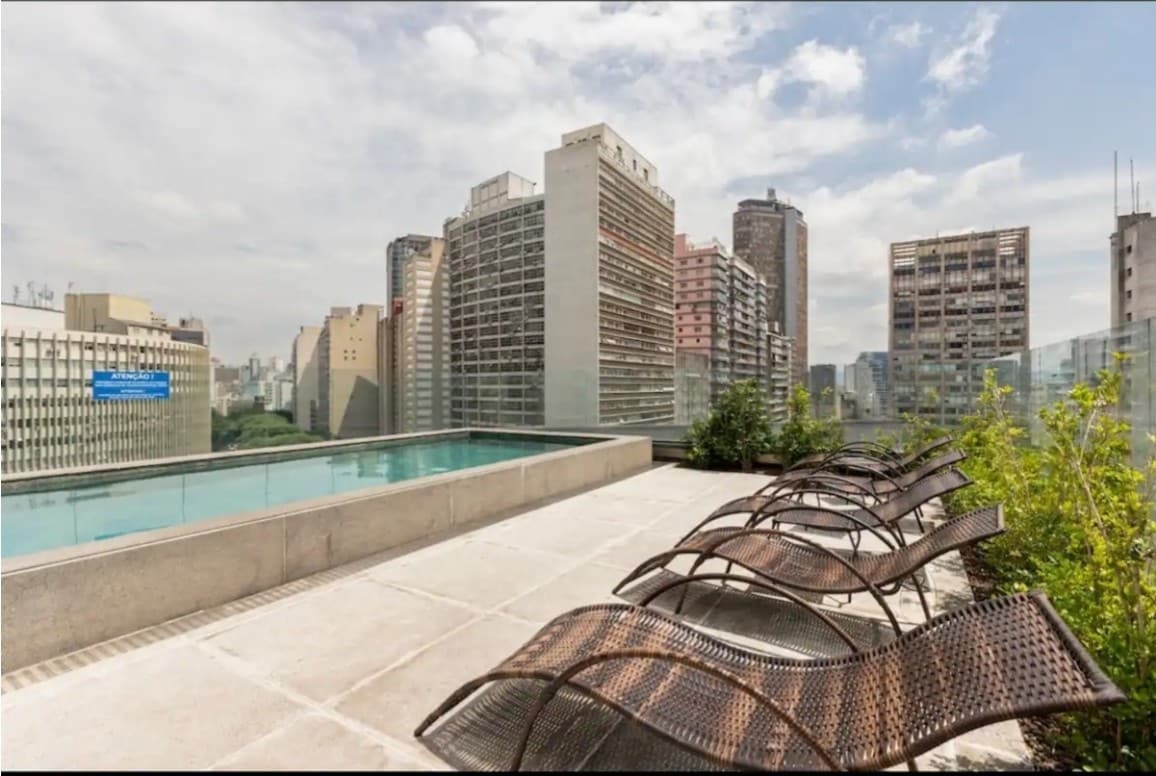 An outdoor pool area is presented with a clear view of the city skyline. Several lounge chairs are positioned to the side of the pool, surrounded by greenery. The sky is filled with clouds, reflecting a bright ambiance.