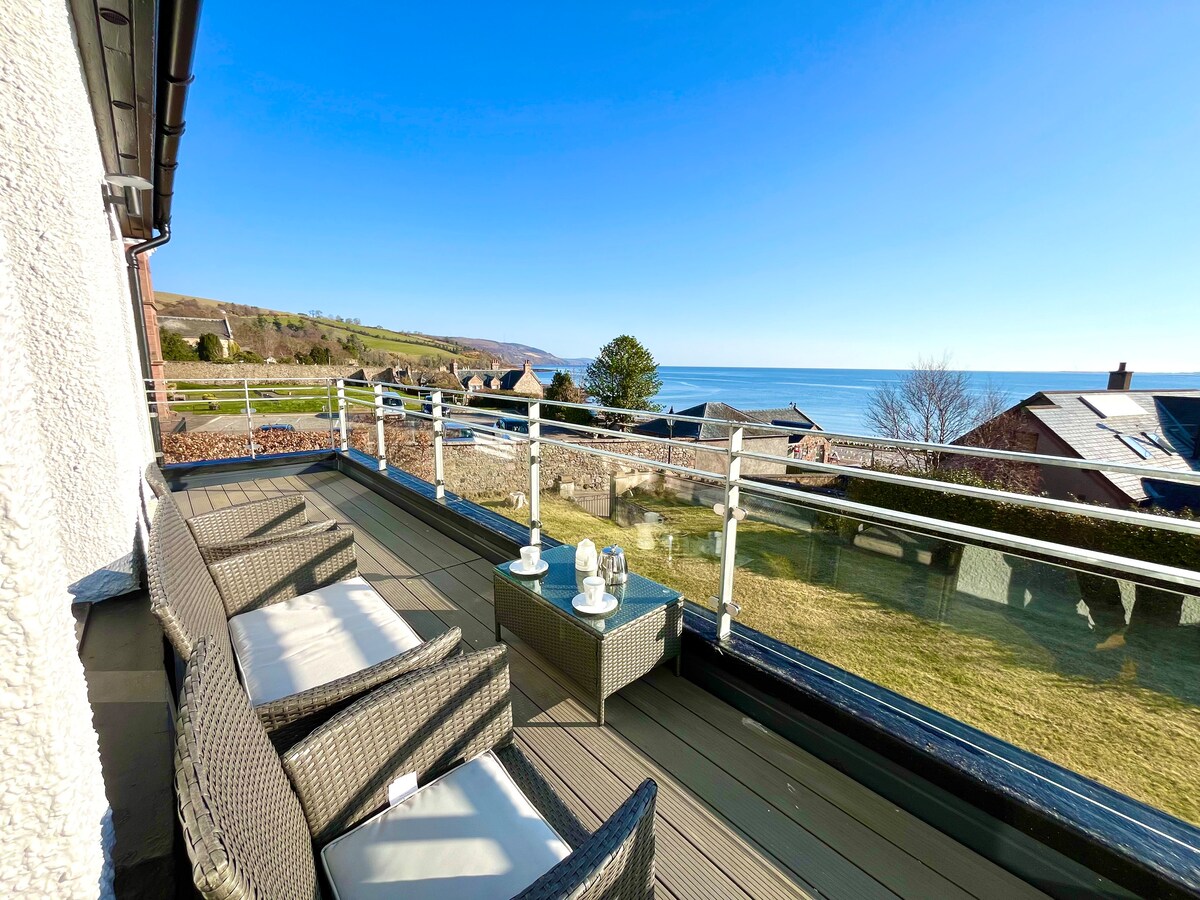 A spacious balcony is presented, featuring comfortable outdoor seating, a glass-top table, and a scenic view of the coastline. The natural surroundings include greenery and distant homes, set against a clear blue sky and the shimmering water of the Moray Firth.