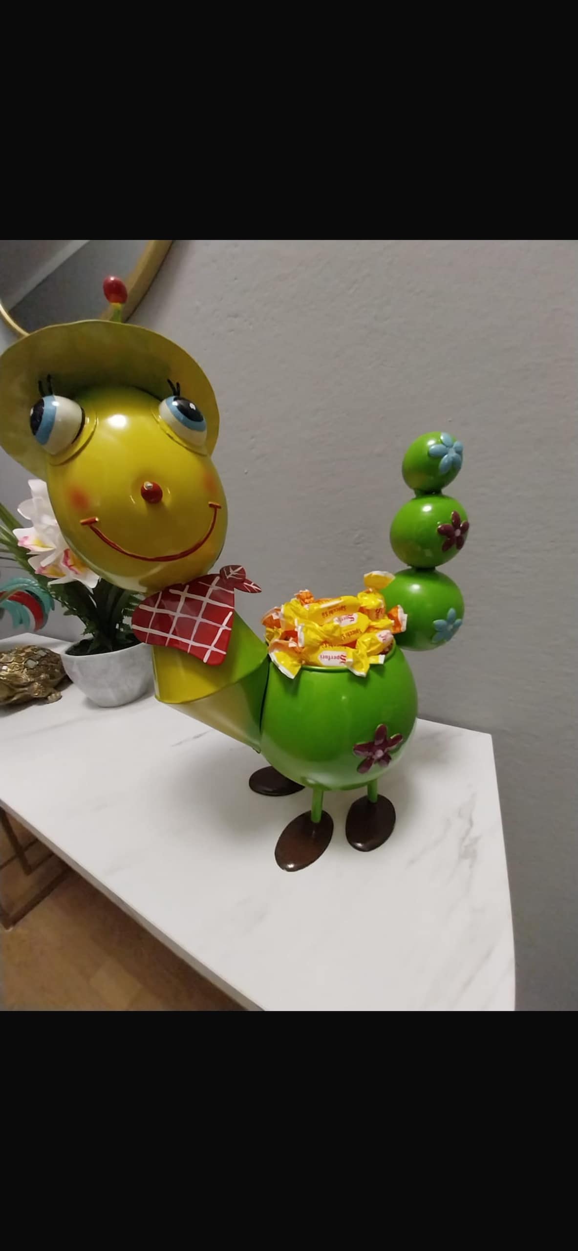 A whimsical decorative piece resembling a cheerful caterpillar is positioned on a marble surface. The caterpillar is bright green with a yellow head, wearing a small hat and a red checkered scarf. Colorful flowers adorn its body, adding a playful touch to the setting.