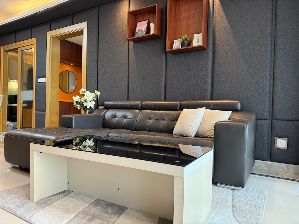 The living room features a modern L-shaped sofa in a dark tone, complemented by a light-colored coffee table with a glossy surface. Decorative shelves are mounted on the wall, showcasing potted plants and framed items. Natural light enhances the open layout.