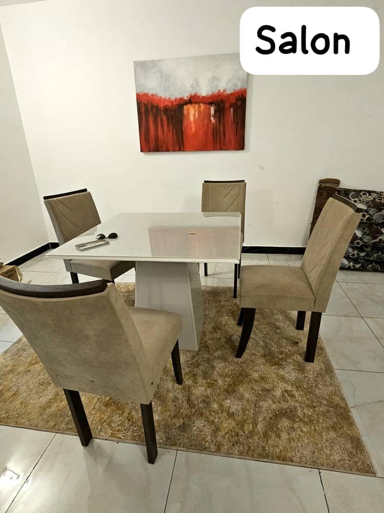 A modern dining area features a square white table surrounded by four upholstered chairs. A soft rug is placed beneath the table, and a large piece of abstract artwork adorns the wall, adding a pop of color to the room.