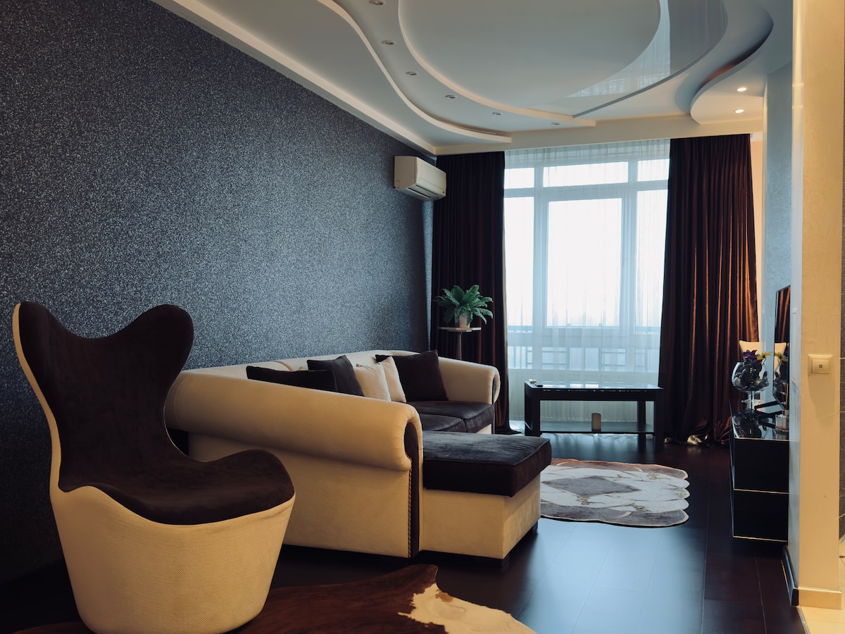 A modern living area features a sleek sectional sofa and a unique sculptural chair positioned on a soft area rug. Large windows allow natural light to fill the space, complementing the textured dark walls and smooth ceiling design. Curtains drape elegantly, adding depth to the atmosphere.