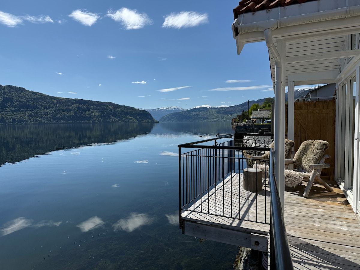 A sunlit deck extends over calm waters, offering views of distant mountains and vibrant blue skies. Comfortable outdoor chairs are positioned along the railing, inviting relaxation while enjoying the serene surroundings. The reflection of the landscape on the water enhances the tranquil atmosphere.