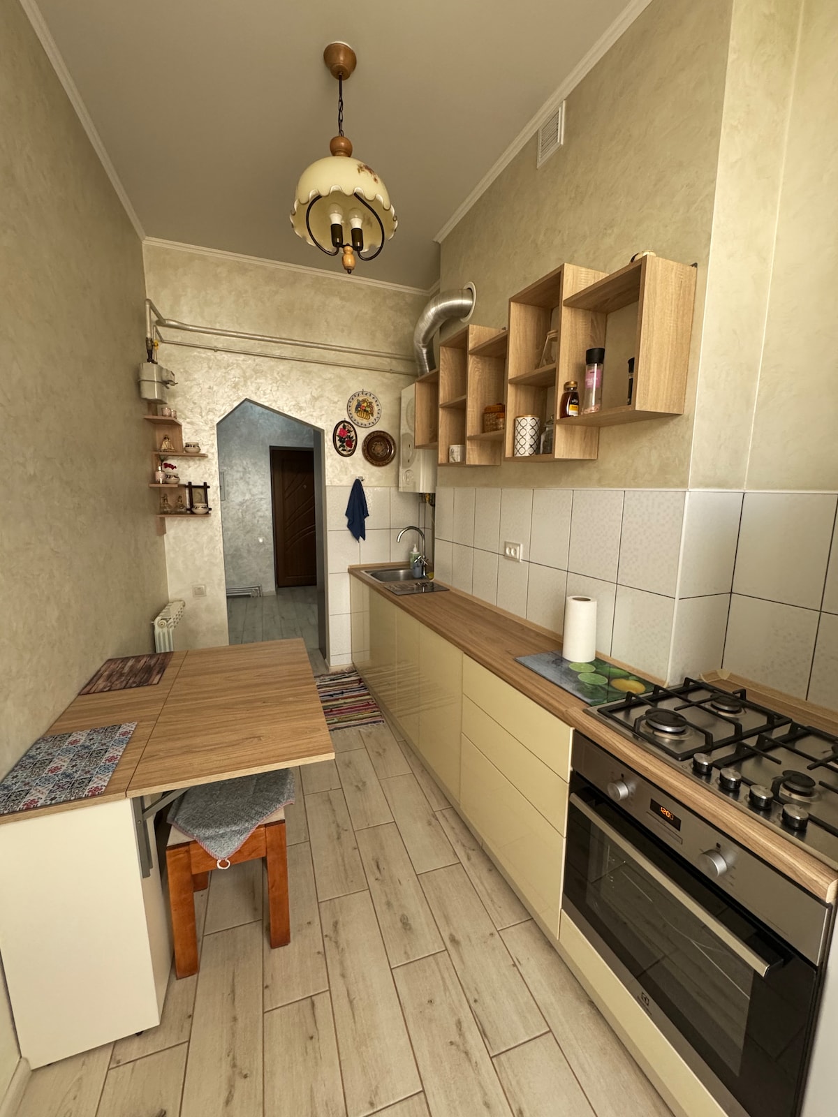 A narrow kitchen space is presented with light-colored cabinetry and a wooden countertop. A small table with a chair is positioned against the wall. Appliances include a gas stove and a sink. Decorative items are displayed on open shelves, adding functional charm to the workspace.