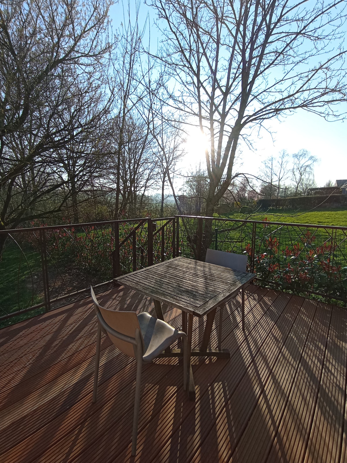 A terrace space is presented, featuring a wooden deck with a small table and two chairs. Sunlight filters through the surrounding trees, illuminating the area while a garden is visible in the background, adding to the serene atmosphere.