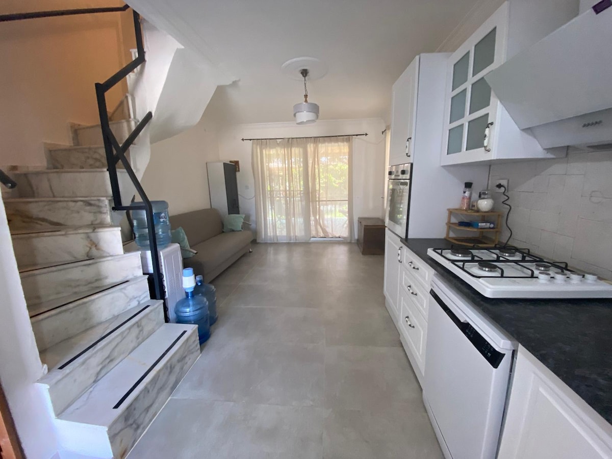 A bright living area is visible, featuring a sofa along one wall and large, draped windows allowing natural light to fill the space. A staircase can be seen leading to another level, while a kitchen corner with a gas stove and cabinets is present on the right.