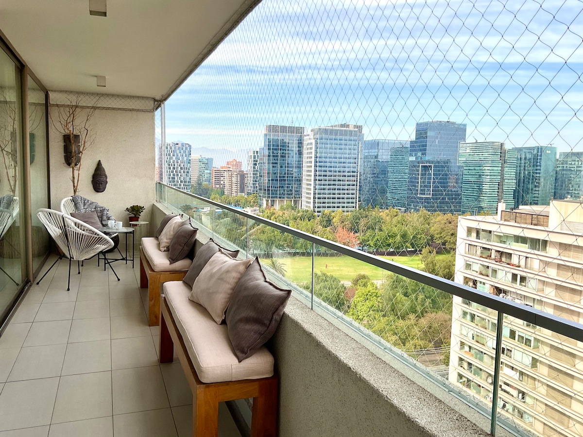 A balcony offers a breathtaking view of a city skyline, with green spaces visible below. Two benches with cushions are positioned along the railing beside potted plants, and two chairs provide additional seating. The area is enclosed with a safety net.