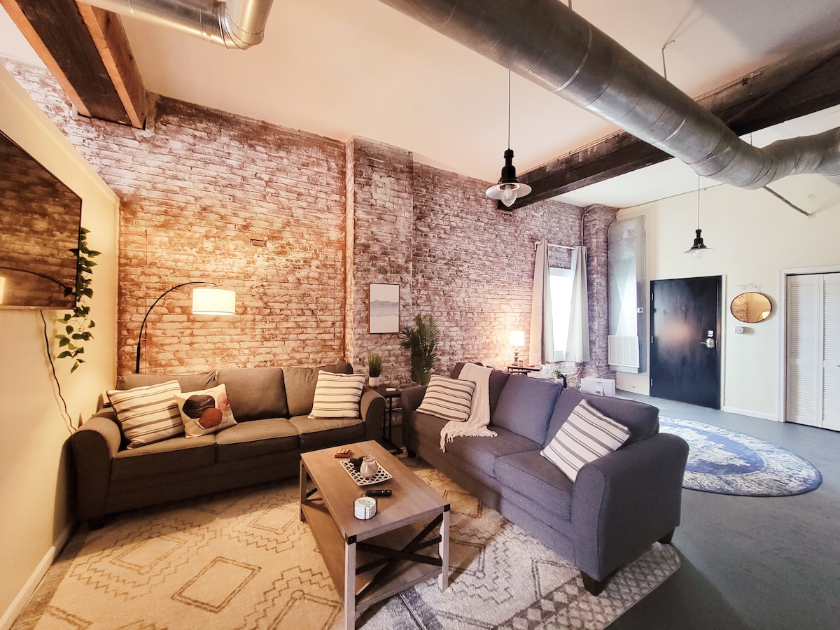 The living area is designed with exposed brick walls and wooden beams. A comfortable gray couch, adorned with striped throw pillows, is complemented by a wooden coffee table. Soft area rugs provide added texture, while large windows allow natural light to illuminate the space.