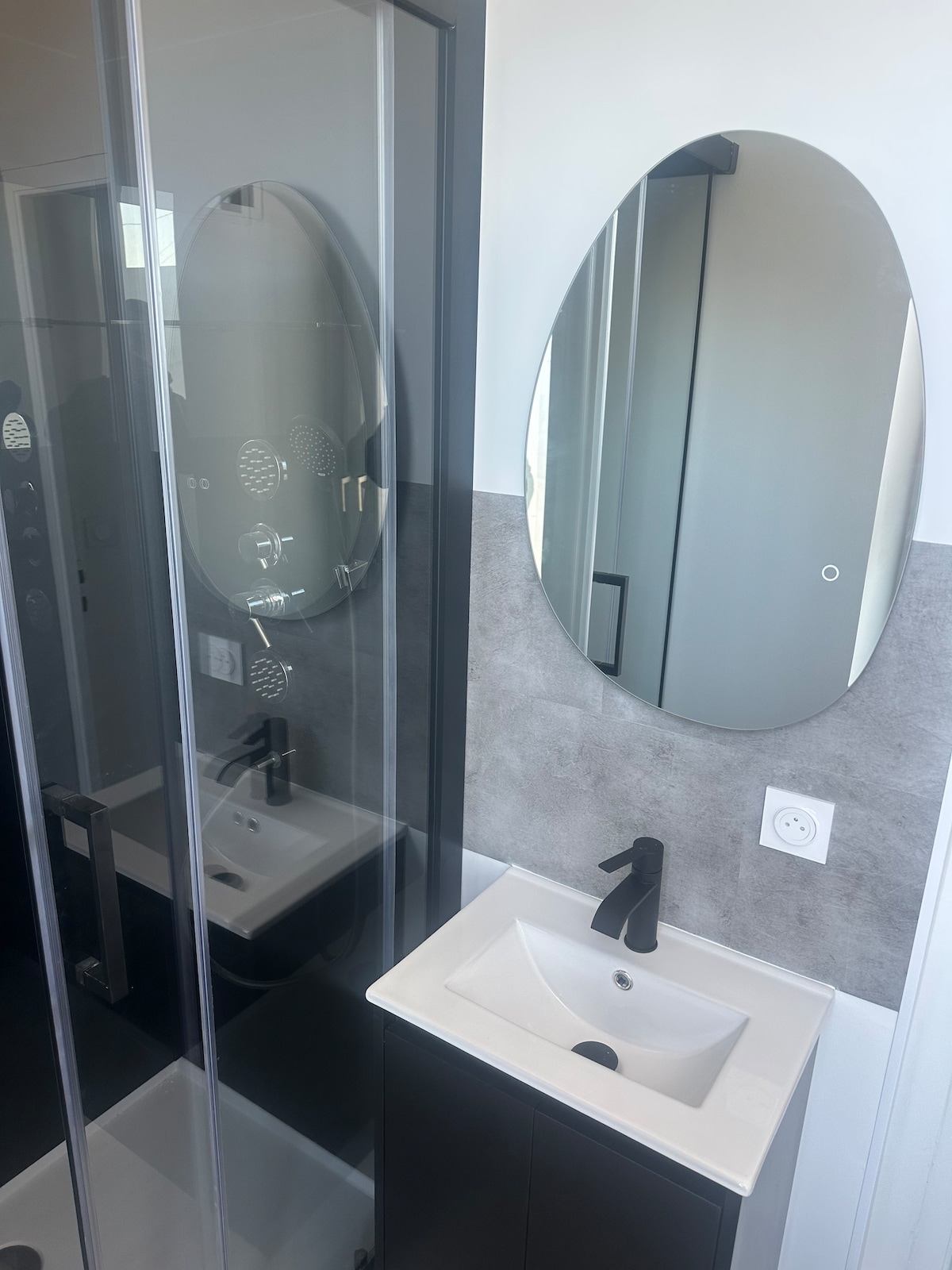A modern bathroom features a sleek, round mirror above a compact sink with a matte black faucet. A glass shower enclosure showcases a minimalist design, while the walls boast a subtle gray finish that complements the space.