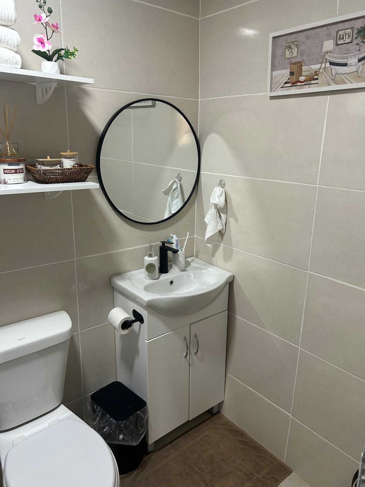 A modern bathroom features light-colored tiles and a round mirror above a compact sink. A small cabinet provides storage beneath the sink, while toiletries and a decorative basket are neatly arranged on a nearby shelf. A towel hangs beside the sink, and a wastebasket is positioned on the floor.