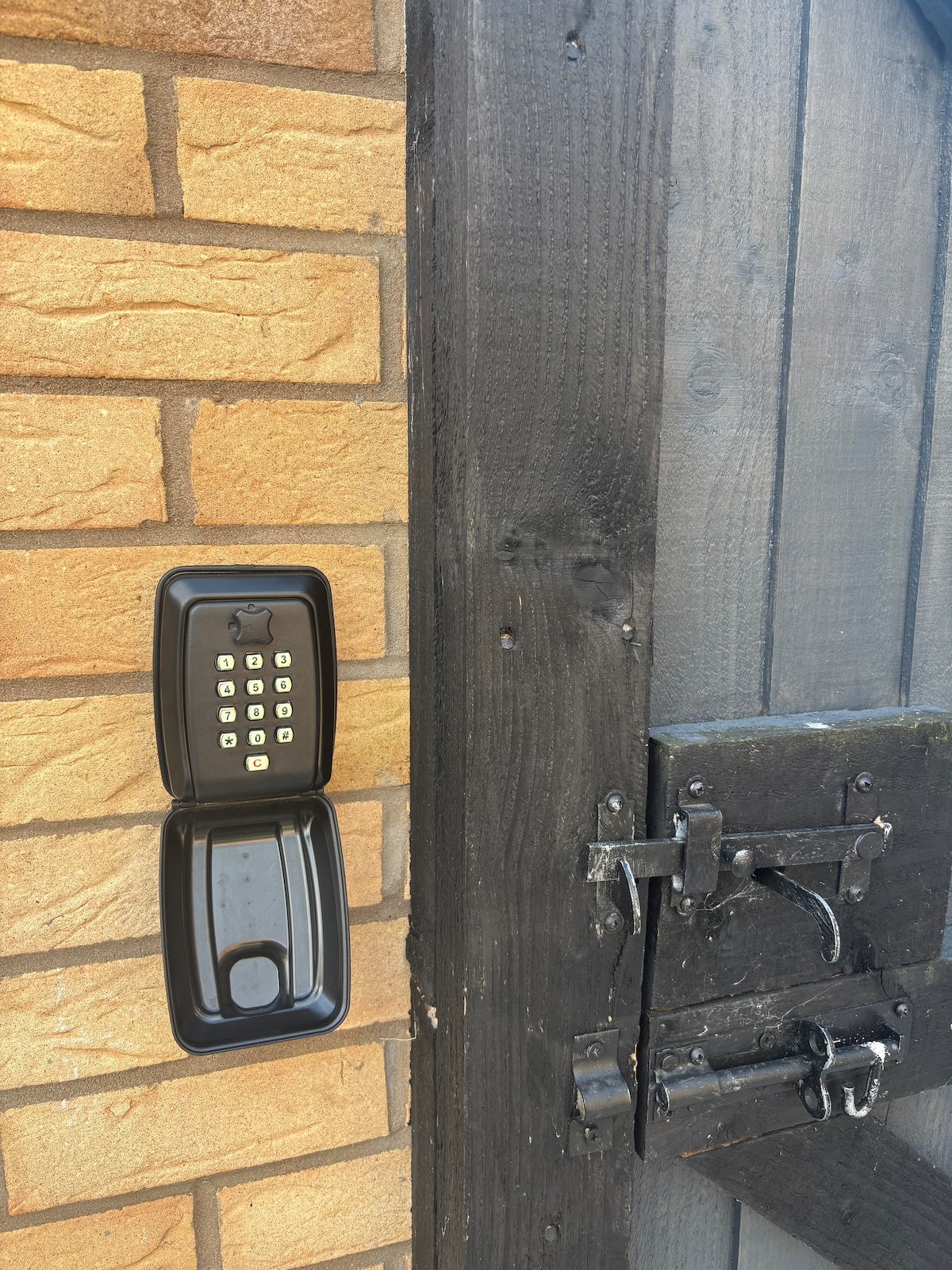 A secure entryway is depicted, featuring a keypad mounted on a brick wall. The keypad is black with clearly visible numbers, positioned beside a locked wooden door. The door showcases a sturdy metal latch and a rustic finish, enhancing the sense of security.