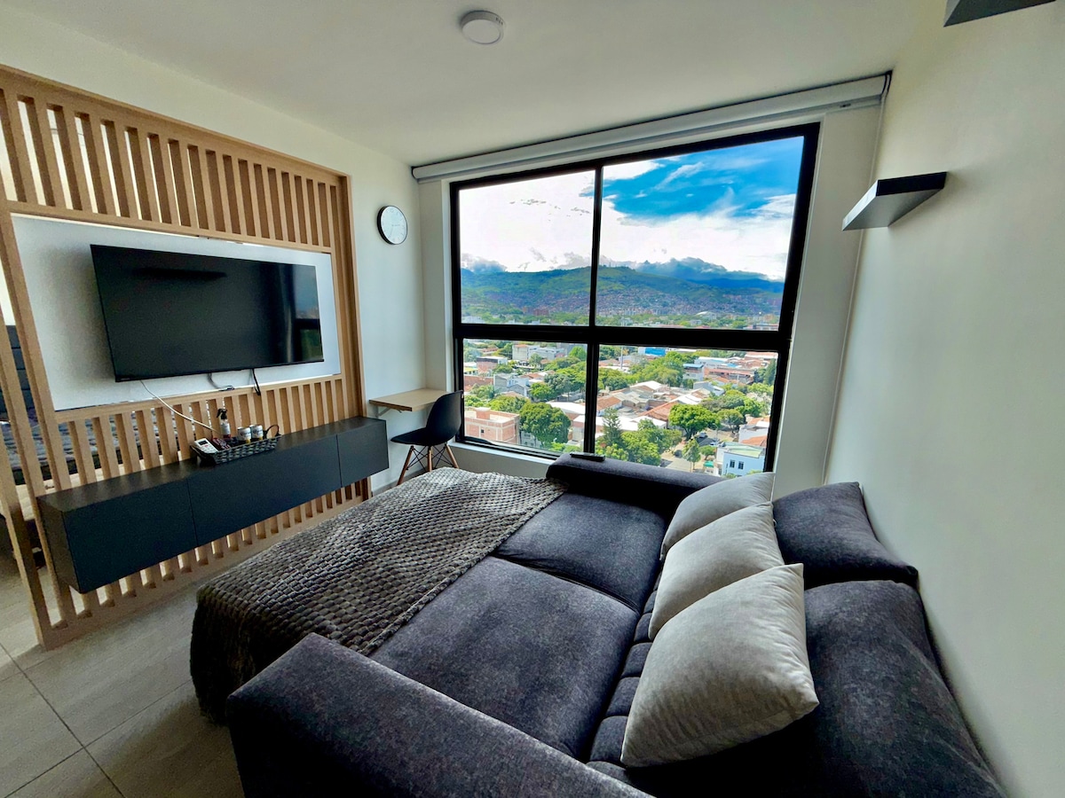 A versatile living area features a dark sofa bed facing a large window, showcasing a view of the city and mountains. A mounted TV sits on a wooden panel alongside a small desk. Natural light fills the space, enhancing the modern design.