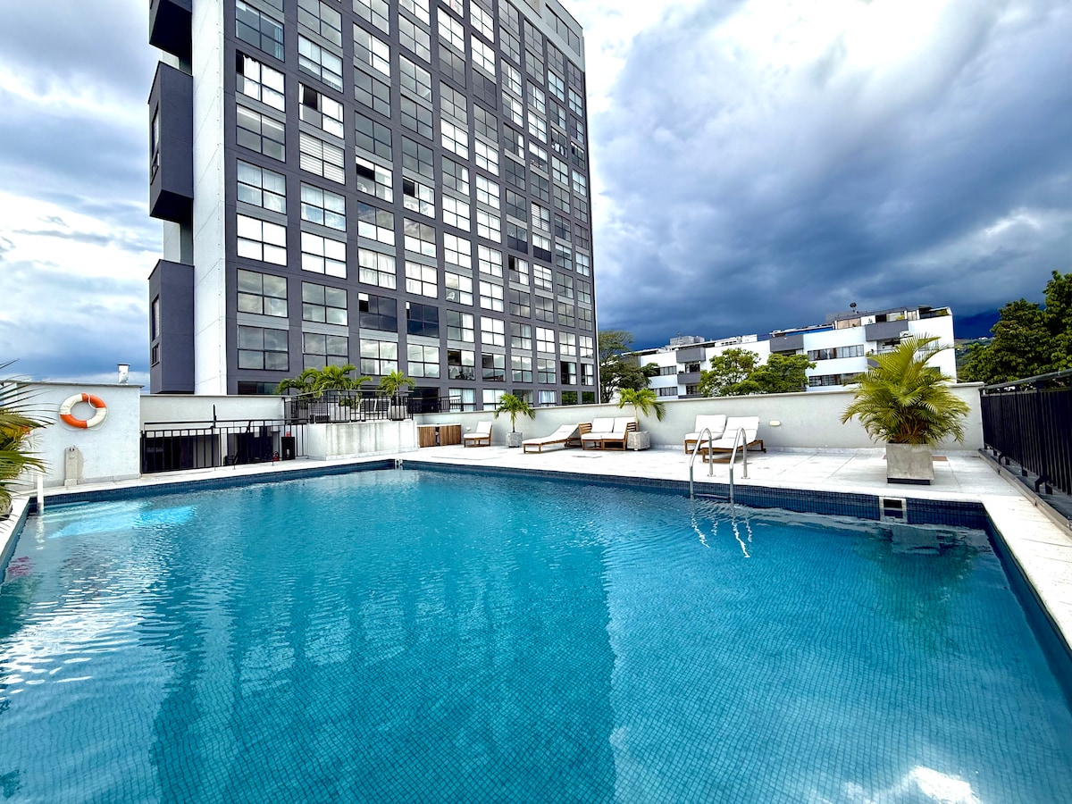 A serene outdoor pool is surrounded by lush palm trees and sun loungers. The building in the background features a modern design with large windows reflecting the sky. The pool's water is clear, inviting relaxation under the clouds.