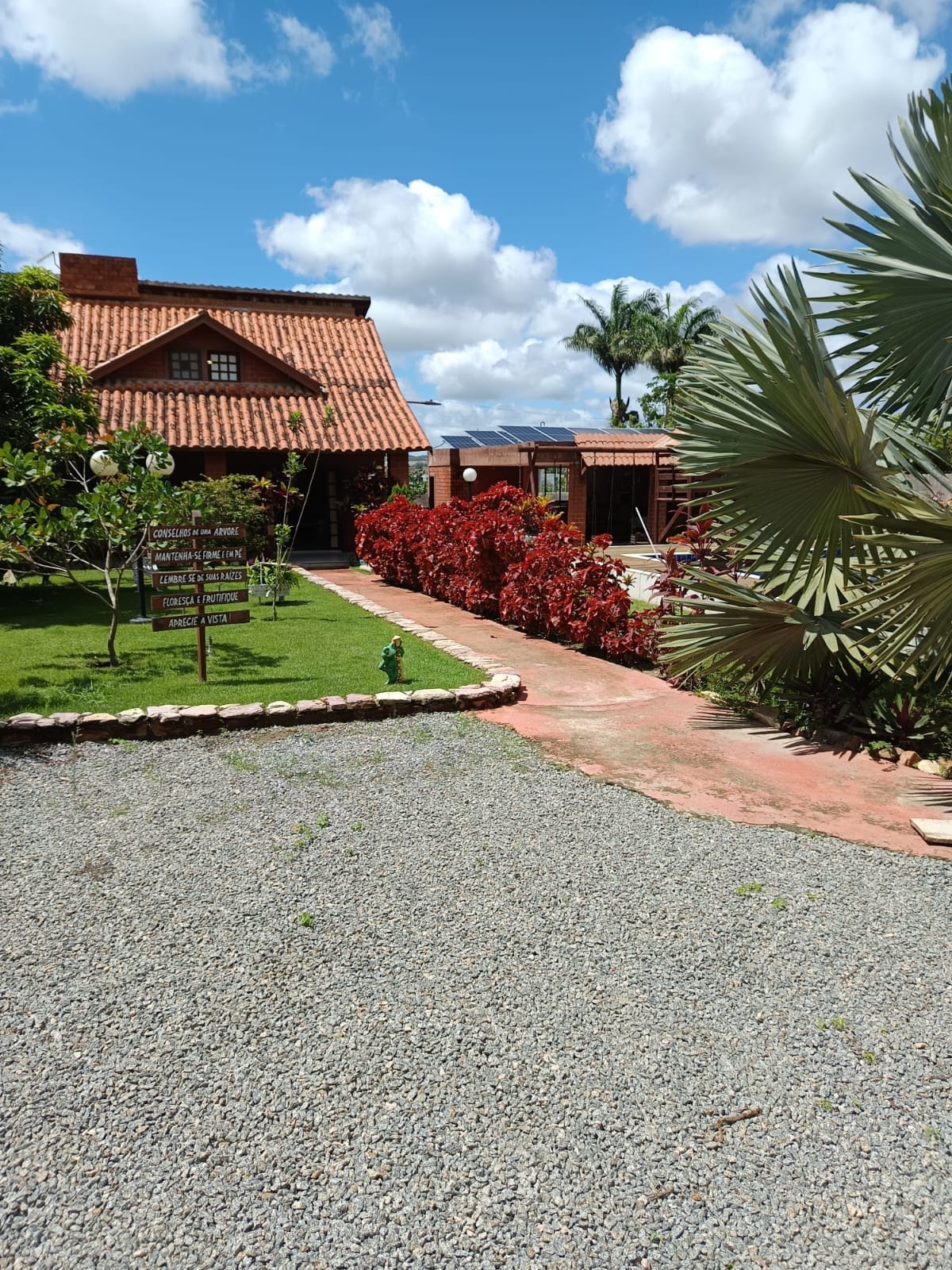 The entrance path leads through a gravel area, flanked by lush greenery and bright red flowering plants. A charming house with a terracotta roof is visible in the background, alongside an outdoor leisure area. The sky is partly cloudy, creating a serene atmosphere.