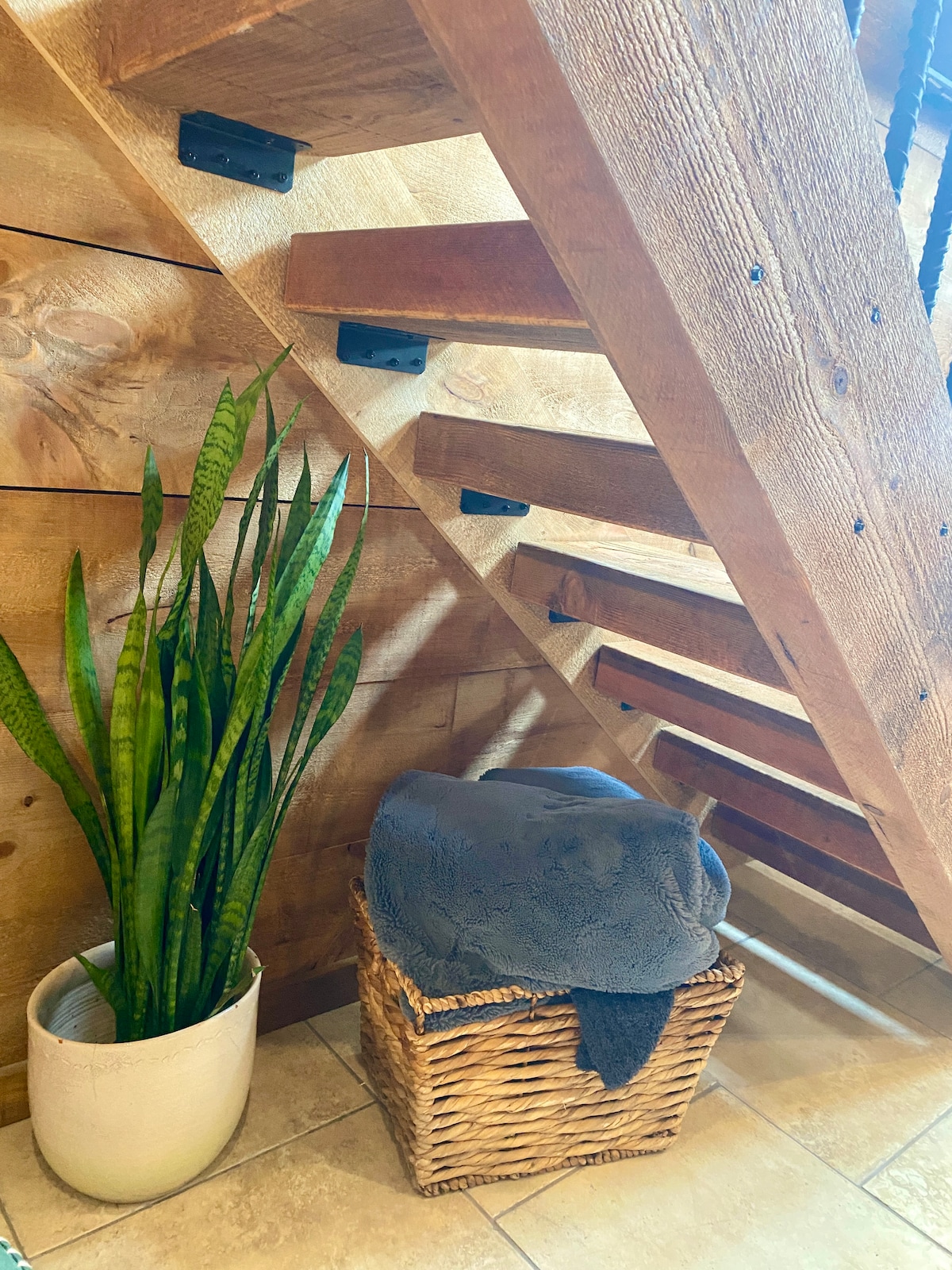 A modern staircase features wooden steps and a wooden wall, creating a warm and natural atmosphere. A tall green plant is placed in a white pot next to a woven basket containing a soft, folded blanket, contributing to the cozy aesthetic.