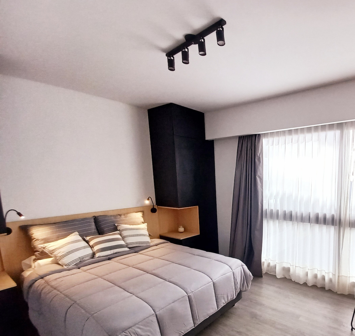 A cozy bedroom features a spacious bed with soft, striped bedding. Ambient lighting is provided by wall-mounted sconces, and large windows are dressed in sheer curtains, allowing natural light to illuminate the room.