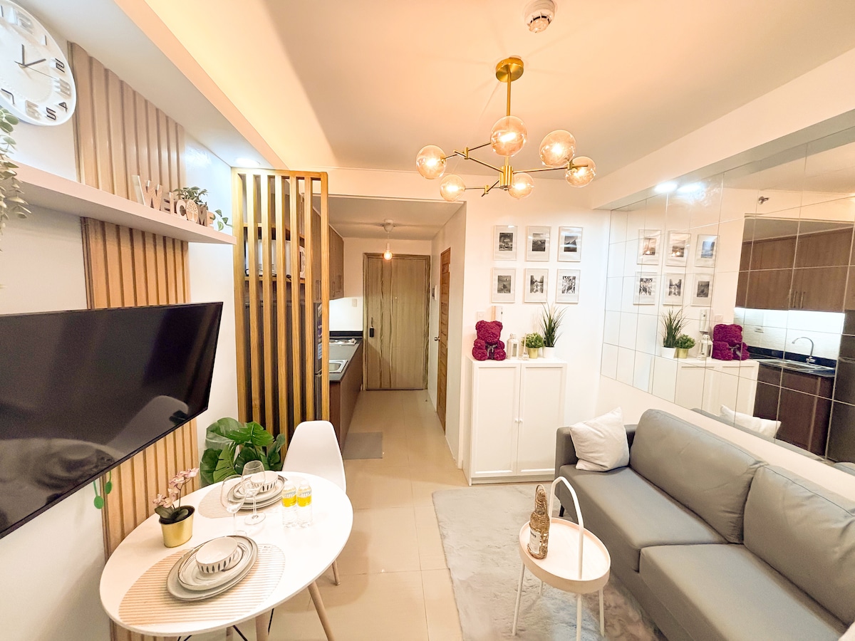 A modern living area features a comfortable sofa positioned beside a small dining set, complete with a circular table and decorative items. Wall art and plants add character, while the space is illuminated by a stylish chandelier. The kitchen area is visible in the background.