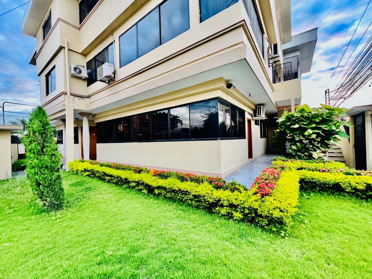 A modern building exterior is captured, featuring large glass windows and a well-maintained garden. Neatly trimmed green grass surrounds vibrant flowering plants, creating a welcoming entrance. Air conditioning units are visible on the side of the structure, enhancing the contemporary design.