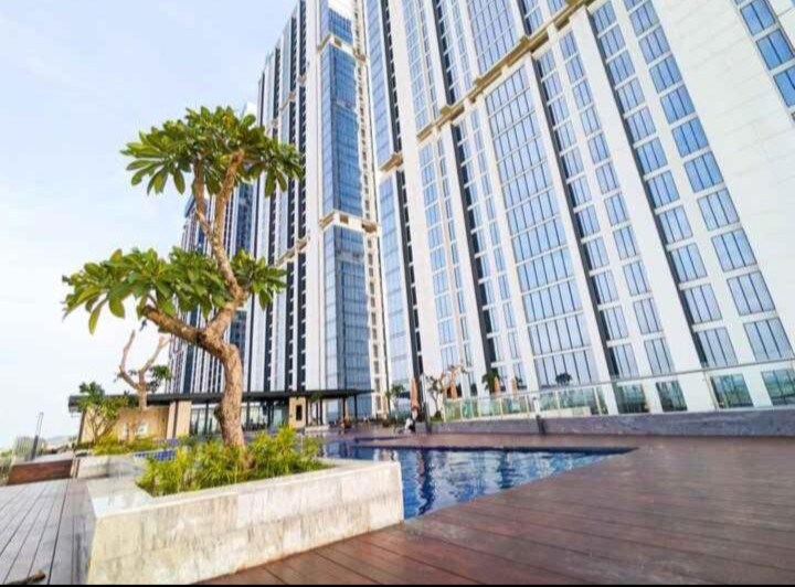 A modern high-rise building features expansive glass windows reflecting the sky. A landscaped area with a pool and a single tree can be seen in the foreground, providing a glimpse of the surrounding outdoor space.