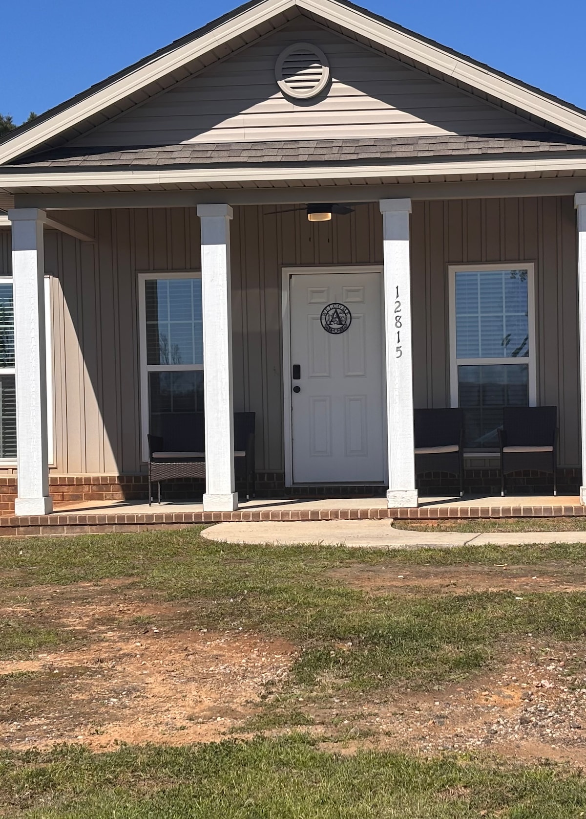The exterior of the home is displayed, featuring a welcoming entrance with a front door flanked by two columns. Two chairs are positioned on either side of the steps, and large windows allow natural light to fill the interior. A clear blue sky serves as the backdrop.