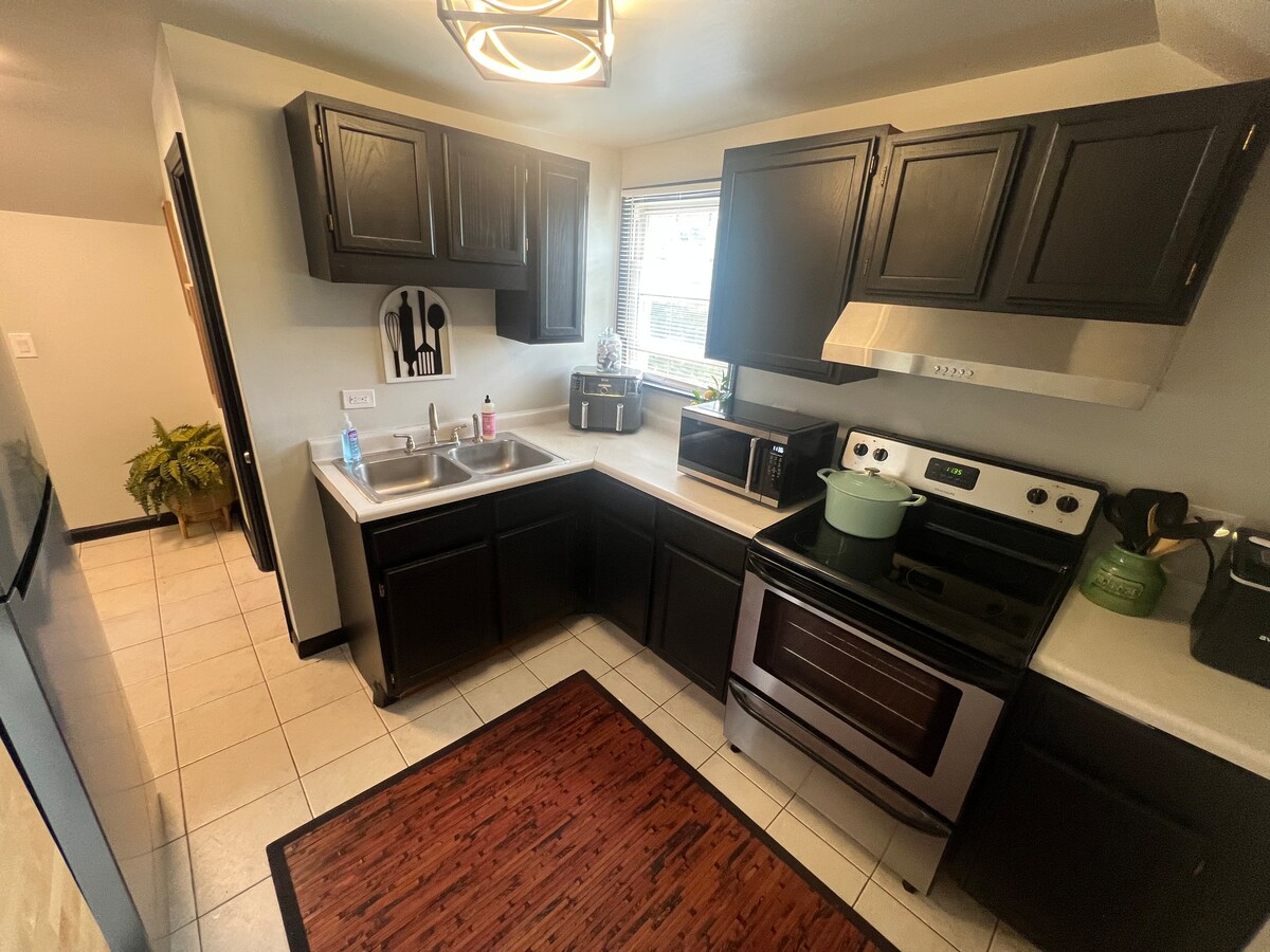 The kitchen features modern dark cabinetry with stainless steel appliances, including a stove and microwave. A spacious countertop extends throughout the area, complemented by a window providing natural light. A rug adds warmth to the tiled floor, enhancing functionality and comfort.