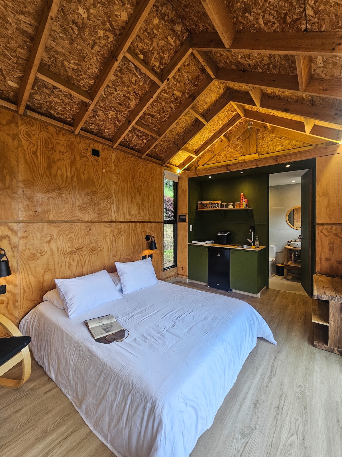 A cozy sleeping area features a spacious bed with a white coverlet, accompanied by a book placed on top. The walls are made of warm-toned wood, and a small desk area is visible in the background, complementing the natural feel of the cabin.