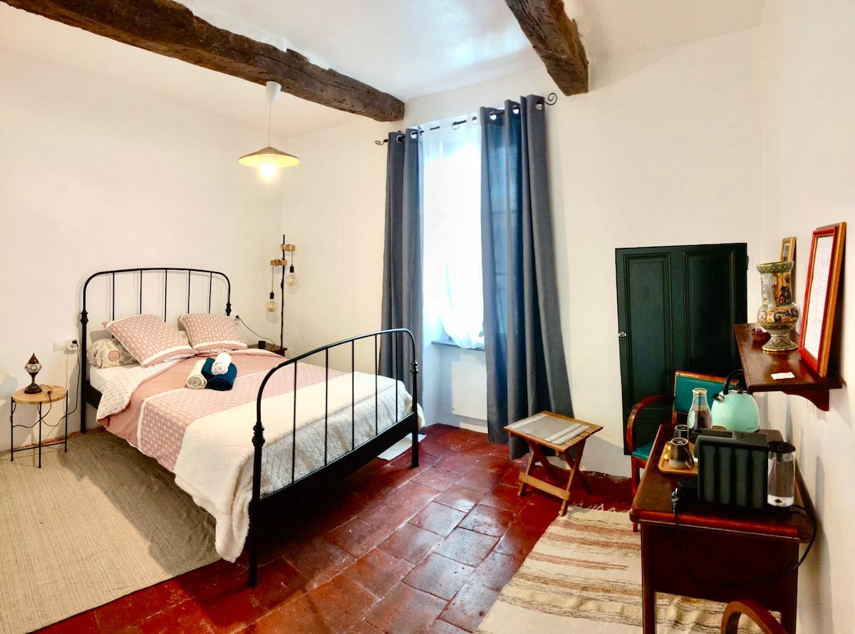 A cozy sleeping area features a double bed with soft bedding and a patterned blanket. Exposed wooden beams highlight the ceiling, while a window with light curtains allows natural light to enter. A small table and a dark cabinet add functional elements to the space.