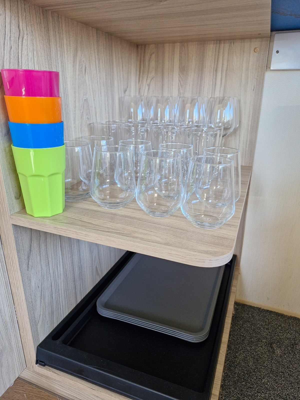 A wooden shelf is organized with colorful plastic cups in vibrant colors stacked at the top. Below, a row of clear glasses is neatly arranged, with a small black tray resting on the shelf underneath. The overall setup emphasizes practicality and accessibility.