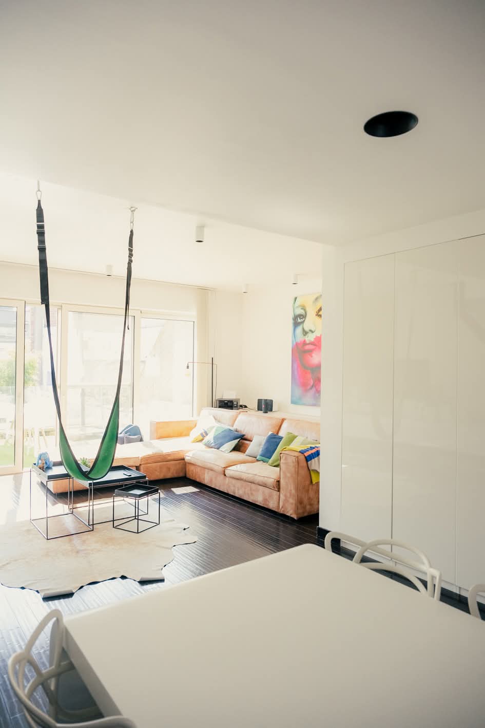 A living area is highlighted with a large, modern sectional sofa adorned with assorted colorful cushions. A suspended green hammock hangs from the ceiling, adding a playful element. Natural light filters through expansive glass doors, illuminating a simple dining table and minimalist decor.