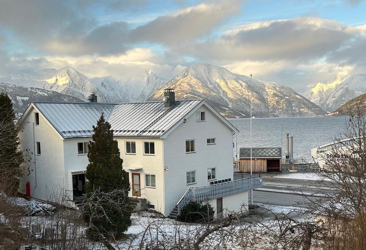 A white multi-story building is positioned near the water's edge, surrounded by snow-capped mountains. A balcony extends along the front, offering views of the fjord. The landscape features a serene winter scene with soft clouds in a blue sky reflecting on the calm water.