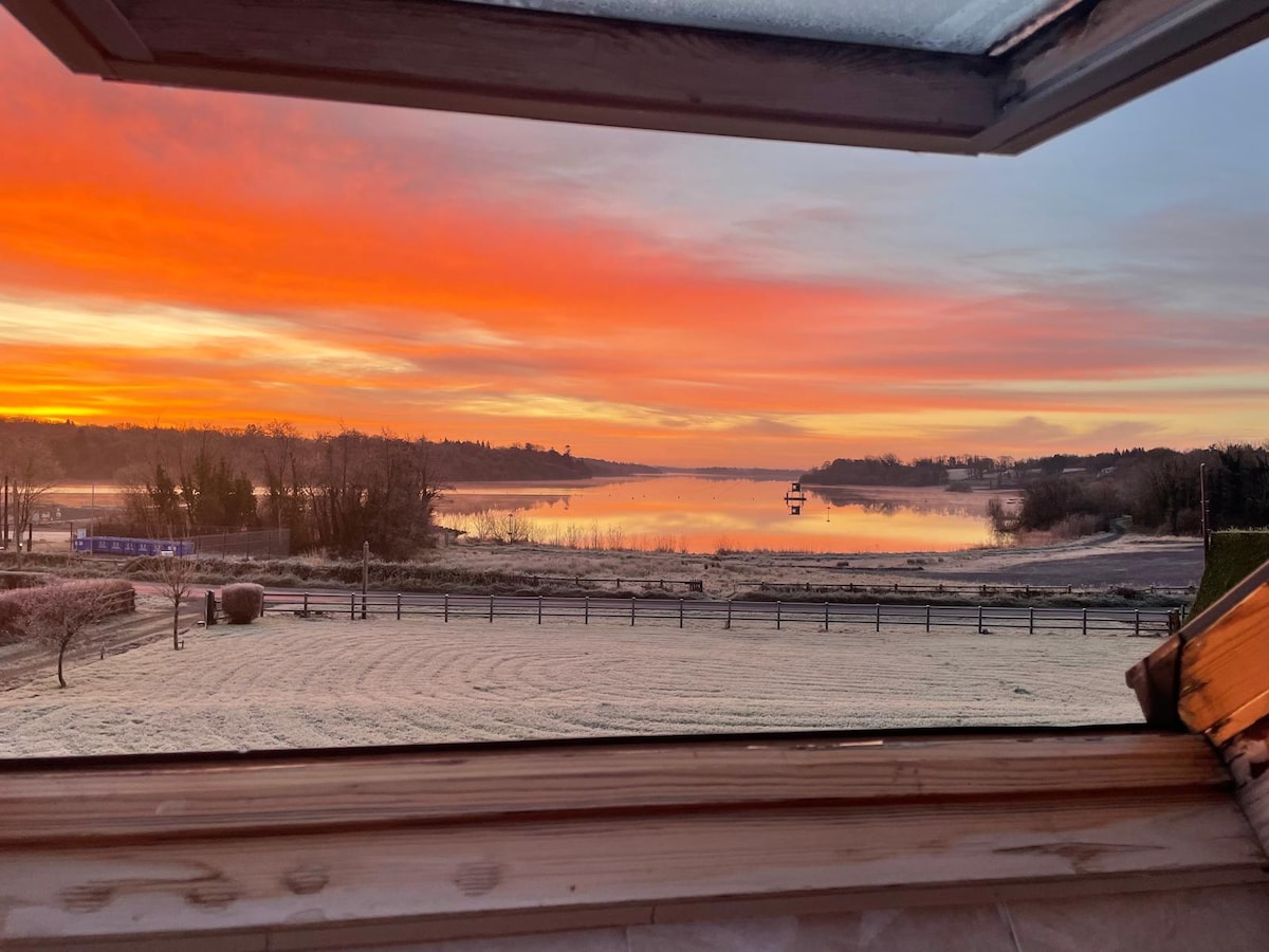 A serene lakeside view is captured through an open window, showcasing vibrant colors of sunrise reflecting on the calm water. The landscape includes frost-covered grass and distant trees, contributing to a peaceful atmosphere.