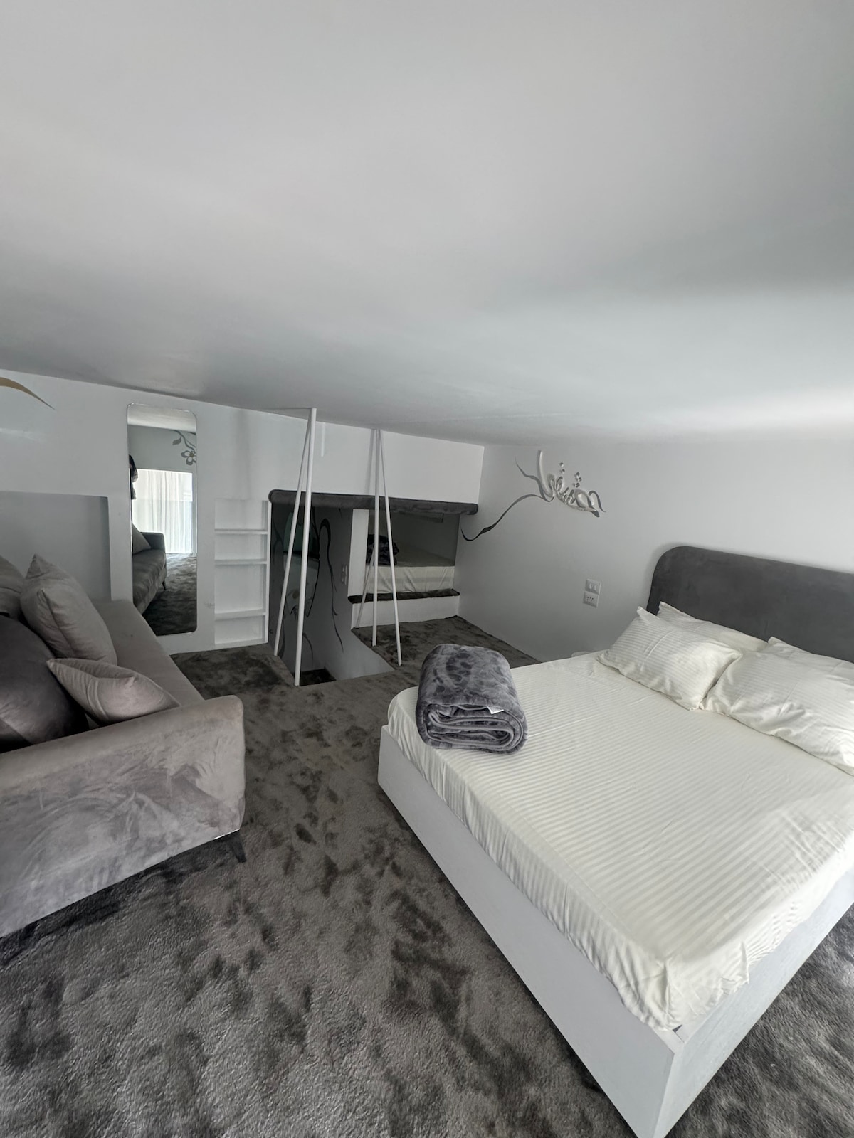 A modern upper floor sleeping area features a spacious double bed with a light-colored mattress. A cozy blanket is neatly folded at the foot of the bed. An accent chair and small shelf are positioned nearby, and natural light filters through a window, illuminating the room.