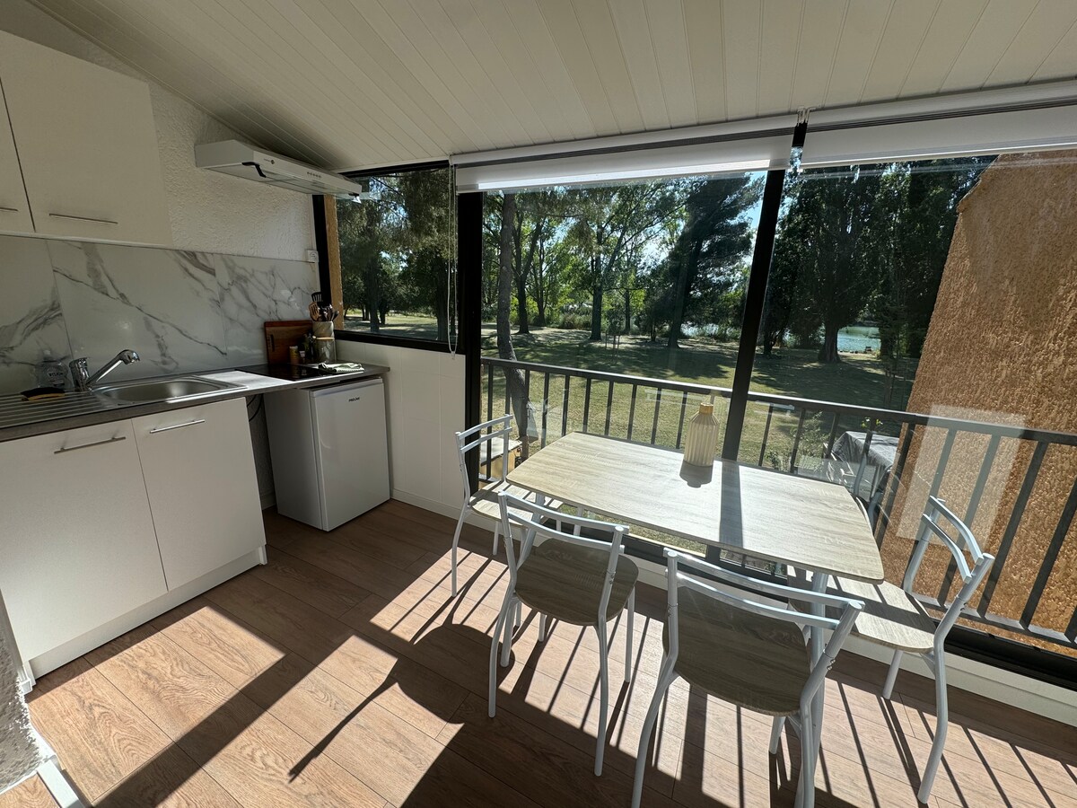A kitchen area is shown with modern white cabinetry and a marble-patterned backsplash. A dining table with four chairs is positioned beside large windows, providing views of a green landscape. Natural light fills the space, enhancing the inviting ambiance of the dining area.