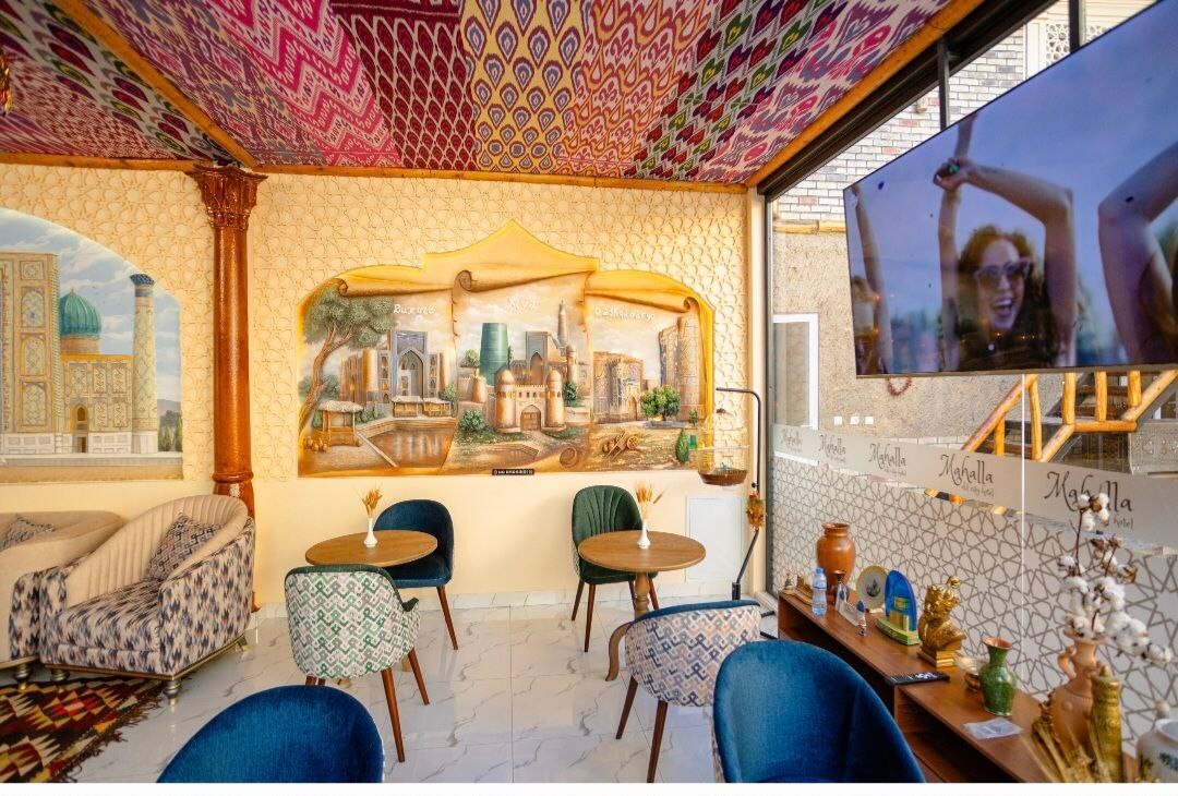 An inviting lounge area is presented with intricately patterned ceiling art and a mural featuring historic architecture. Two small tables are surrounded by upholstered chairs, complementing the vibrant decor. A TV screen is mounted on the wall, enhancing the communal atmosphere.