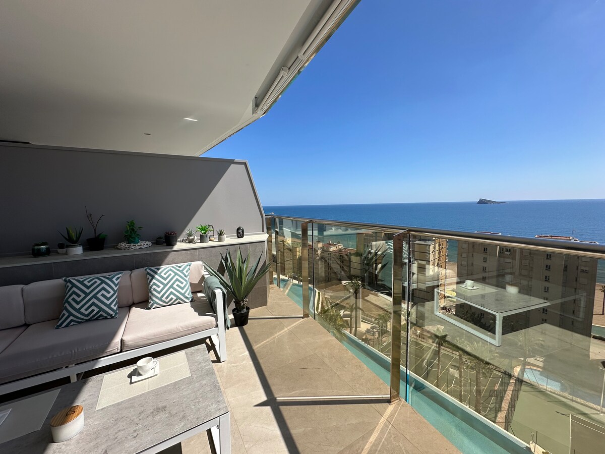 A spacious terrace features a comfortable seating area with soft cushions, surrounded by green plants. The panoramic view reveals the serene Mediterranean Sea, accentuated by clear blue skies. Glass railings provide an unobstructed sightline, ideal for enjoying the outdoors.