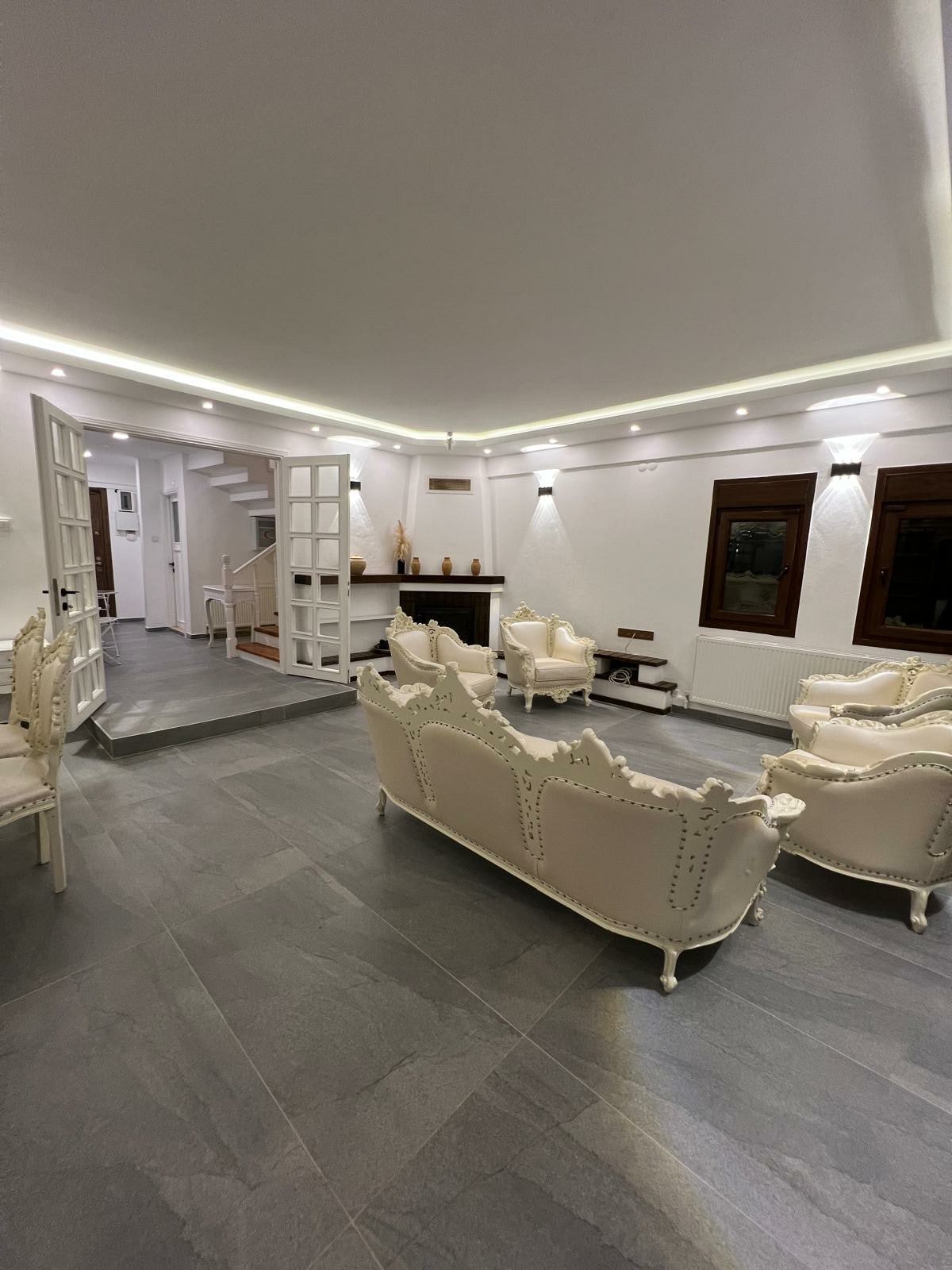 A bright living area is presented with a soft color palette. Elegant white seating is arranged for comfort, featuring decorative detailing. The space is enhanced by indirect lighting, and a staircase is visible in the background, providing access to upper levels.