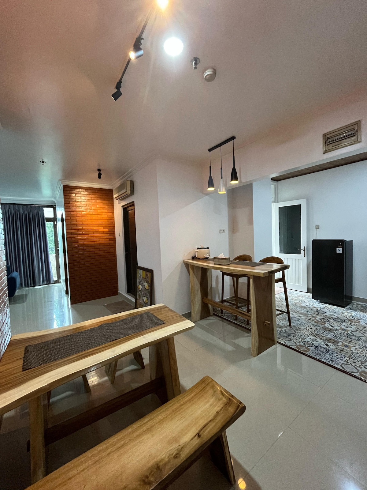 A modern dining area features a wooden table accompanied by two benches. Pendant lights hang above, providing ambient illumination. A stylish rug lies beneath the table, with a small refrigerator and a door visible in the background. Windows offer a glimpse of natural light.