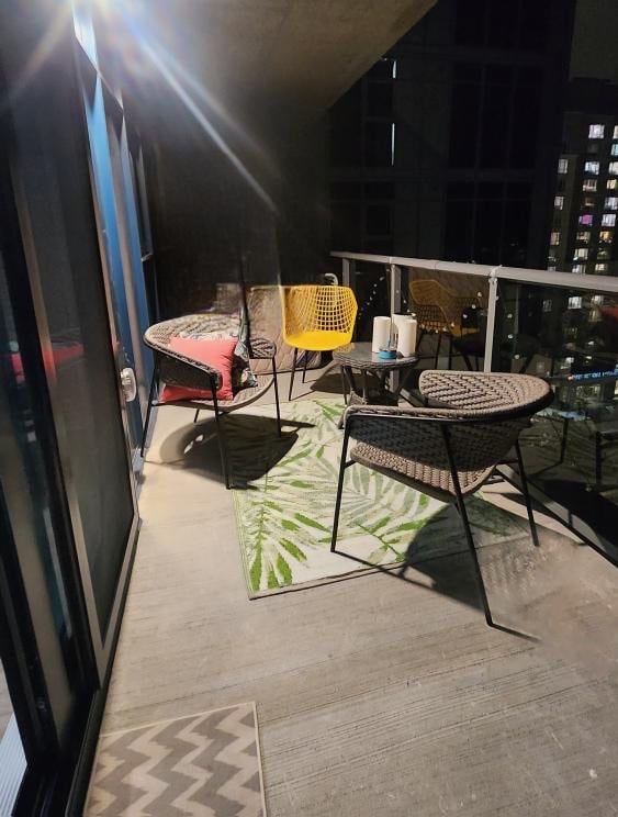 An evening balcony is furnished with four chairs: two black woven chairs and two yellow accent chairs. A small table sits in the center, surrounded by a decorative rug featuring green leaves. Soft lighting enhances the serene outdoor setting against a backdrop of city lights.