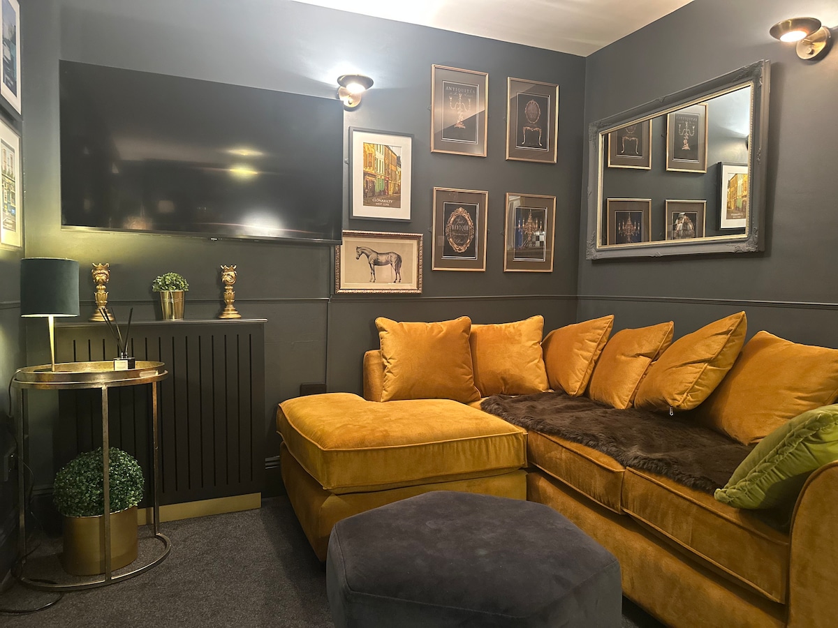 A cozy living area features a plush yellow sectional sofa with coordinating throw pillows. A circular ottoman rests nearby on a carpeted floor. Wall-mounted lighting highlights an array of framed artwork, and a flat-screen television is positioned above a sleek, decorative console.