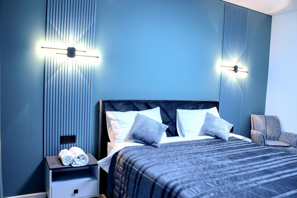 A stylish bedroom features a deep blue accent wall, complemented by soft lighting. A large bed is adorned with a textured gray blanket, flanked by plush pillows. A cozy armchair is positioned in the corner, while neatly rolled towels rest on a bedside table.