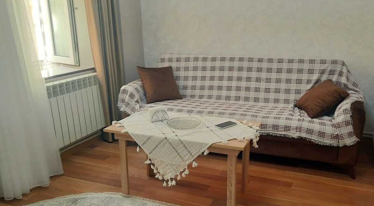 A living room area is showcased featuring a cozy sofa covered with a checked throw. A small wooden table is positioned in front of the sofa, adorned with a decorative tablecloth and a few items. Natural light filters in through a nearby window with soft curtains.
