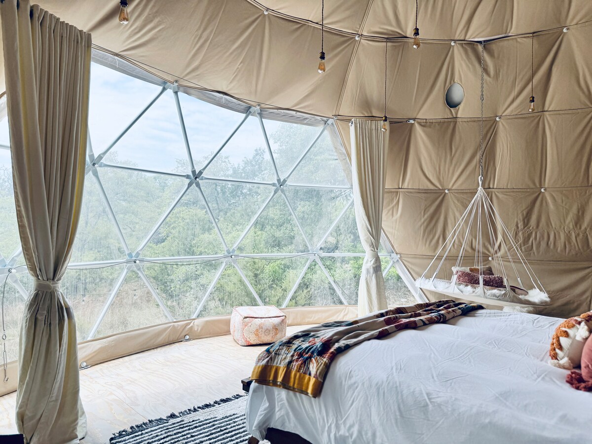 A cozy bedroom is highlighted by a spacious bed adorned with soft bedding. A wall of windows allows natural light to fill the room, revealing views of the surrounding landscape. A hammock is suspended beside the bed, adding a unique touch to the serene space.
