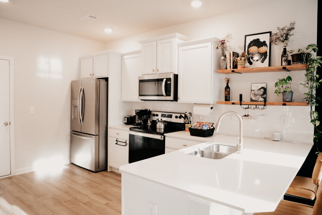 A modern kitchen is designed with white cabinetry and a contrasting black stove. Stainless steel appliances are integrated, while open shelving displays decorative items and potted plants. A large island with a sink provides additional workspace, complemented by ample overhead lighting.