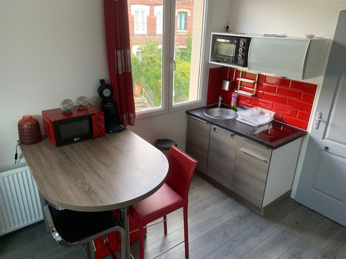 The kitchen area includes a dining table with two red chairs and a compact kitchenette. Modern appliances are visible, such as a microwave and coffee maker. Bright red backsplash tiles contrast with the light cabinetry, while natural light enters through a window overlooking greenery.