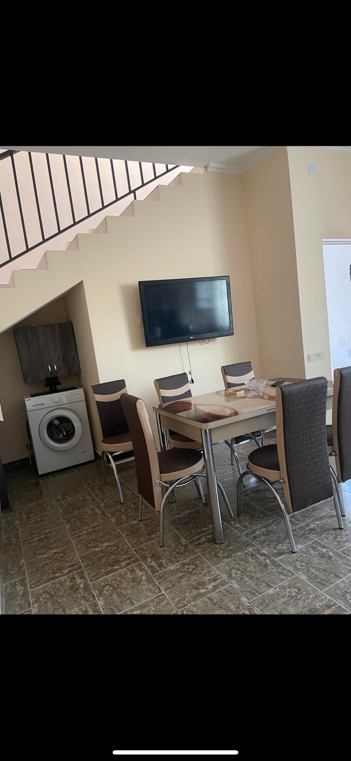 A dining area is featured, equipped with a table and six chairs. A flat-screen television is mounted on the wall, and a washing machine is positioned nearby. The space benefits from natural light, highlighting the tiled flooring and neutral wall colors.